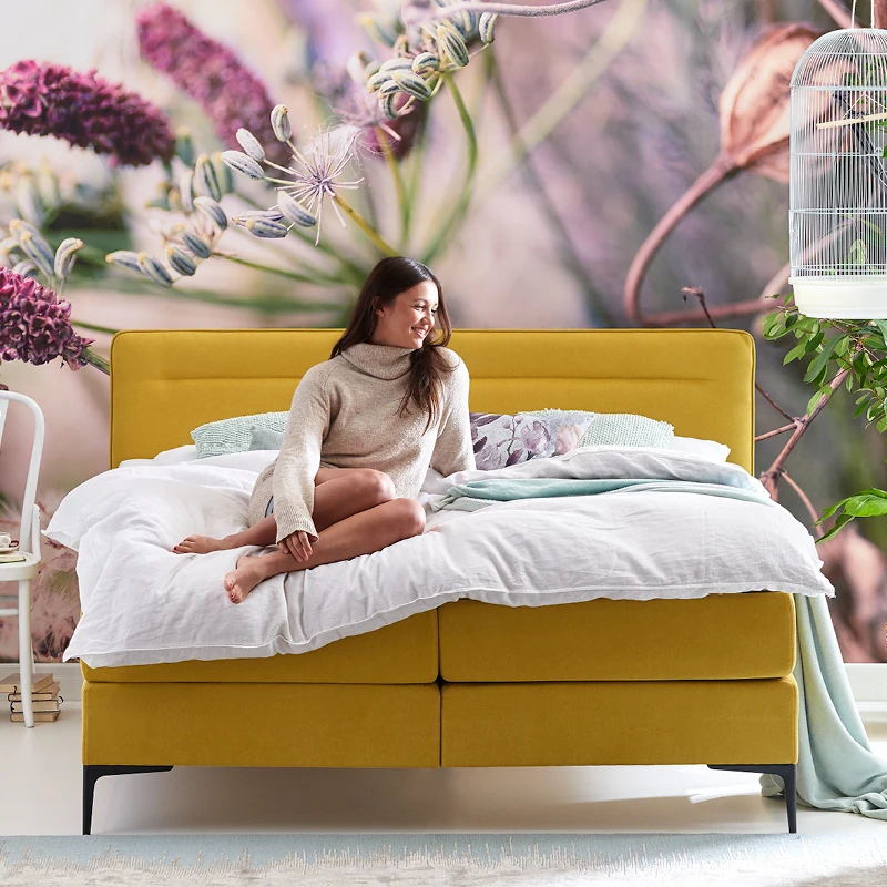 Boxspring Tender in mosterdgeel, met een laag hoofdbord en luchtig wit beddengoed